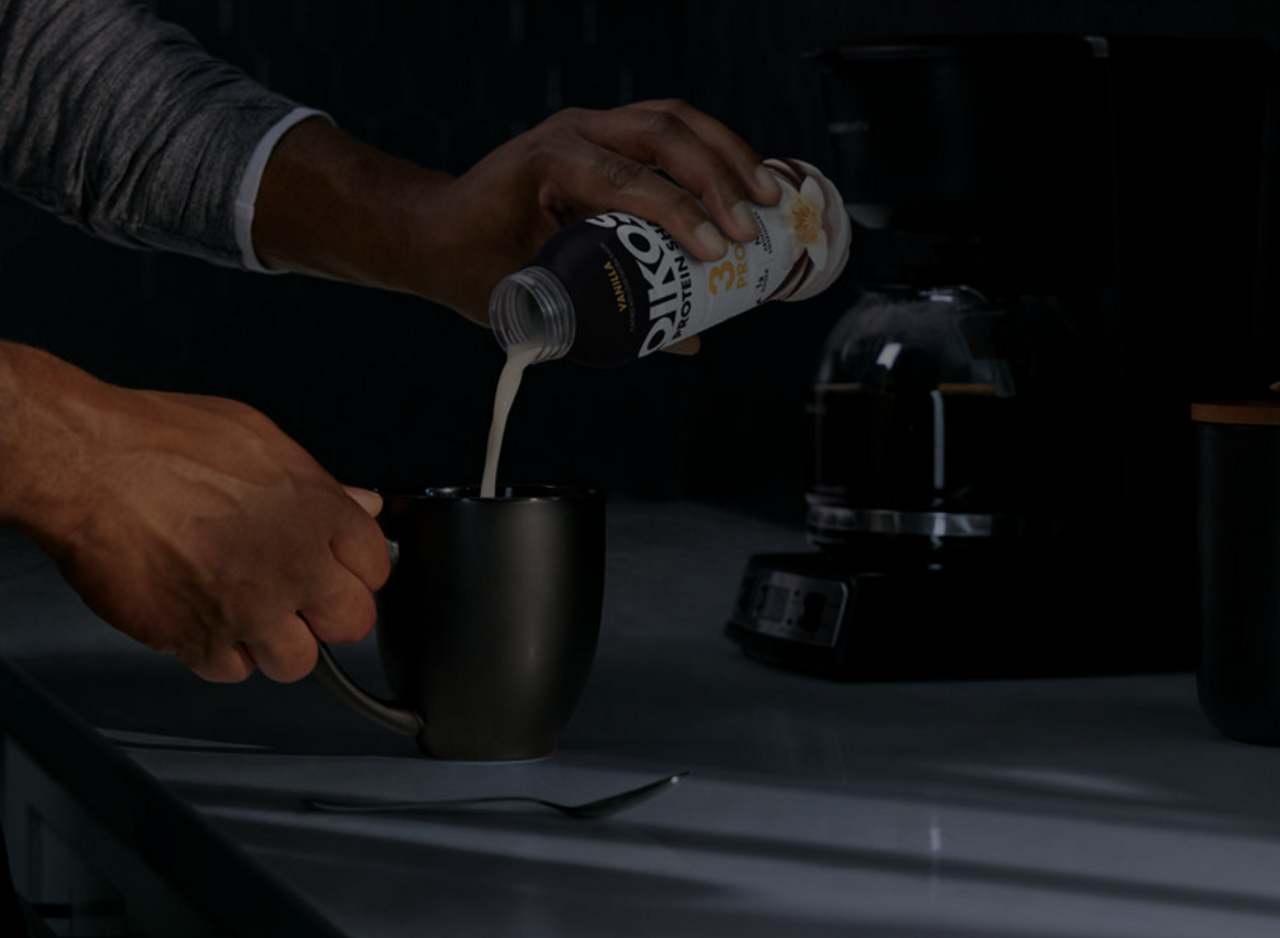 A person pours from a Oikos Protein Shake bottle into a black coffee mug.