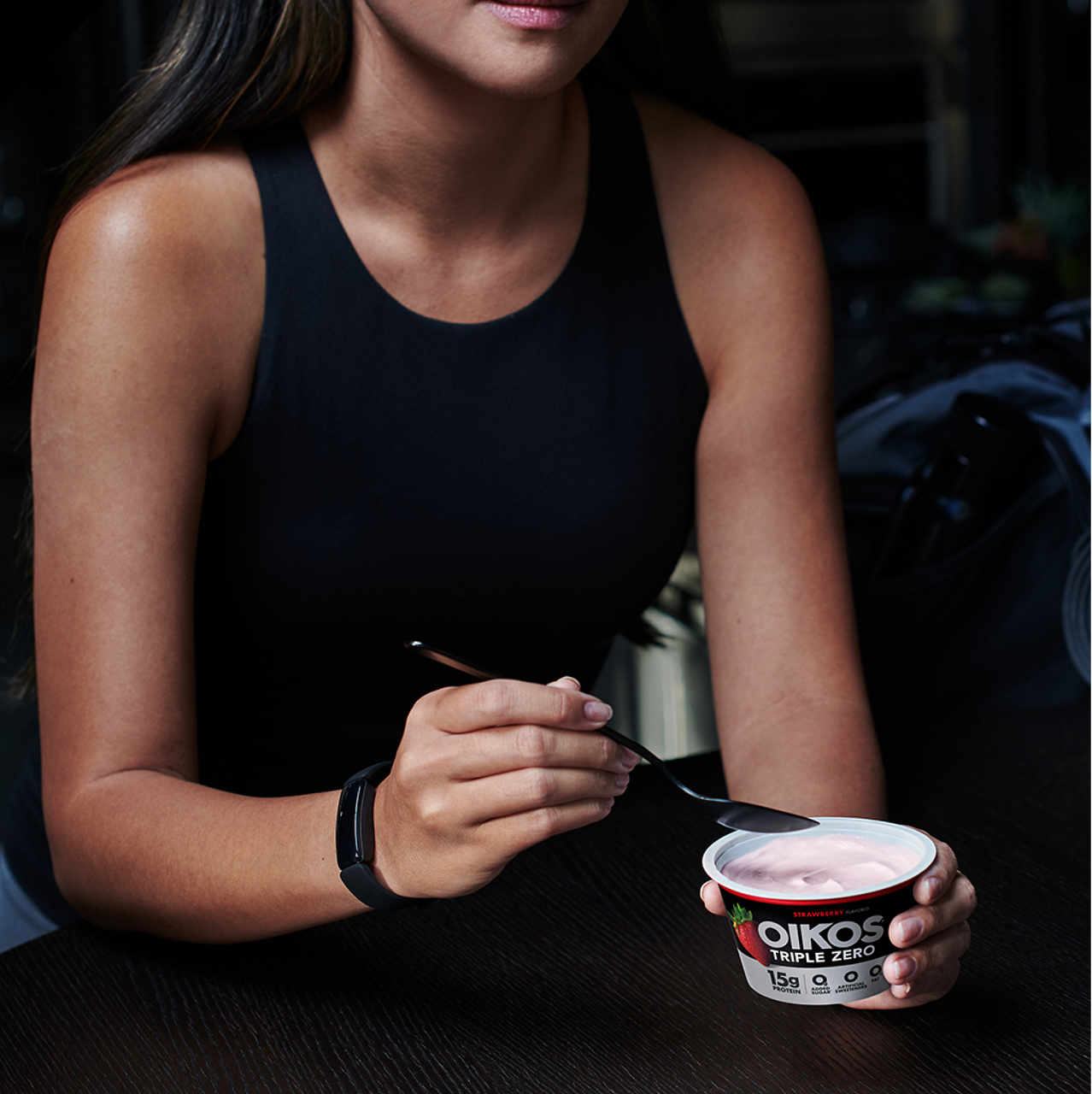 A young woman in a black sleeveless top holds a spoon and a cup of Oikos Triple Zero yogurt. 